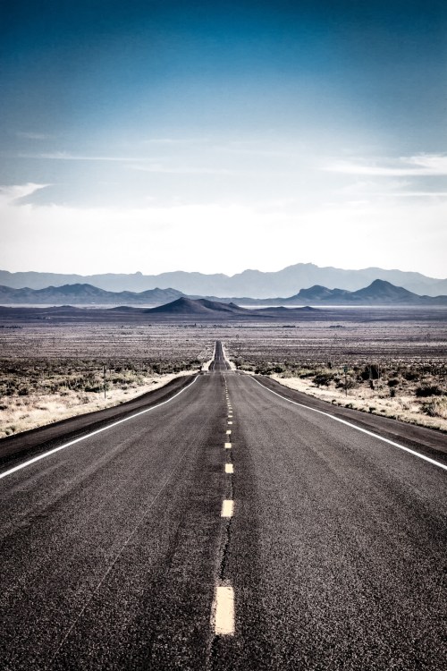A long, empty road stretches into the distance, flanked by desert landscape and mountains under a clear blue sky.