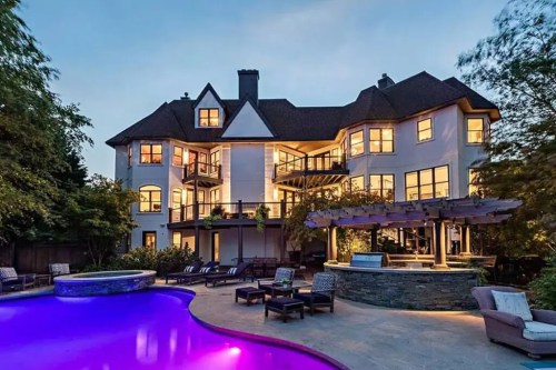 A luxurious mansion with large windows, a pool, and outdoor seating, illuminated at dusk.
