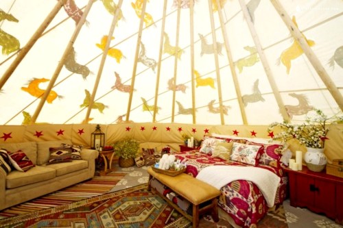 Cozy interior of a teepee with a bed, sofa, and colorful decorations, featuring patterned textiles and warm lighting.