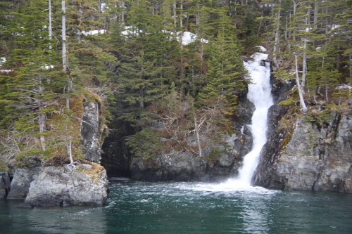 A serene waterfall cascades down rocky cliffs into a tranquil, greenish-blue body of water, surrounded by evergreen trees.