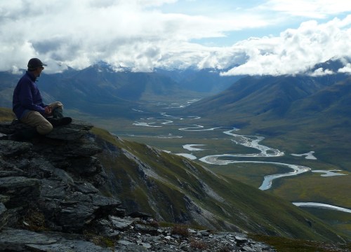 A person sits on a rocky ledge, overlooking a winding river and expansive valley under a cloudy sky.