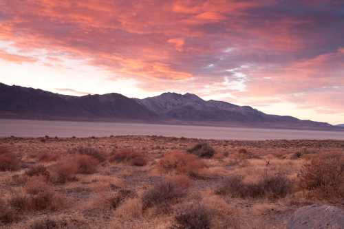 A desert landscape at sunset, featuring mountains in the background and colorful clouds in the sky.