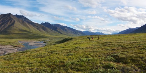 Three hikers explore a lush green valley surrounded by mountains under a blue sky with scattered clouds.