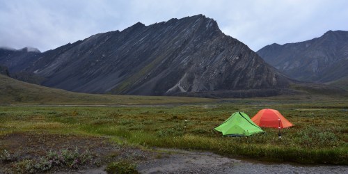 Two tents, one green and one orange, set against a rugged mountain landscape under a cloudy sky.