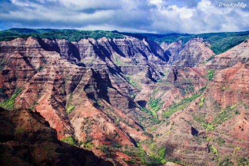 A panoramic view of colorful, rugged canyons with lush greenery and dramatic clouds in the sky.