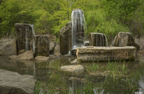A serene water feature with cascading waterfalls over large rocks, surrounded by greenery and a calm pond.