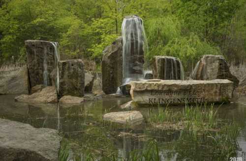 A serene water feature with cascading waterfalls over large rocks, surrounded by greenery and a calm pond.