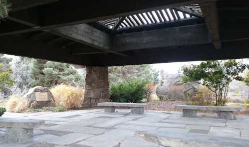 A shaded stone seating area with benches, surrounded by greenery and rocks, under a wooden structure.