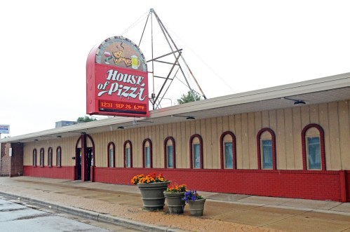 Exterior of "House of Pizza" restaurant with a red sign, flower pots, and a cloudy sky in the background.