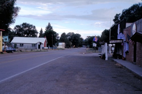A quiet street scene in a small town, featuring buildings, trees, and a clear sky.