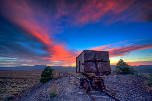 An old train car sits on abandoned tracks under a vibrant sunset with colorful clouds and distant mountains.