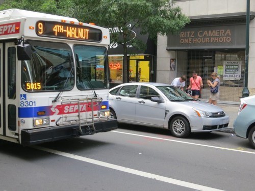 A SEPTA bus on a city street near a camera shop, with pedestrians and a parked silver car in the background.
