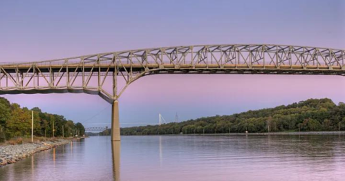 A large metal bridge spans a calm river, surrounded by trees under a pastel sky at sunset.