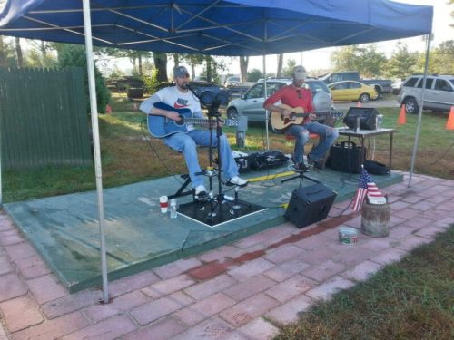 Two musicians play acoustic guitars under a blue canopy, with a small audience and parked cars in the background.