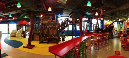 Indoor play area featuring colorful slides, climbing structures, and palm tree decorations. Brightly lit with playful seating.