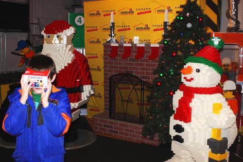 A child holds a camera in front of LEGO sculptures of Santa and a snowman, with a decorated Christmas tree nearby.
