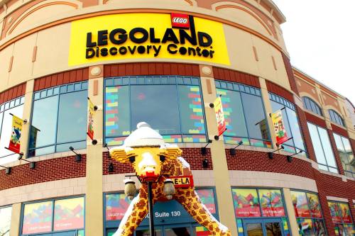 Exterior of the LEGOLAND Discovery Center, featuring a large LEGO giraffe sculpture and colorful banners.