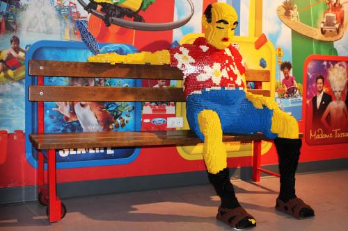 A colorful LEGO sculpture of a man in a Hawaiian shirt sitting on a bench against a vibrant background.