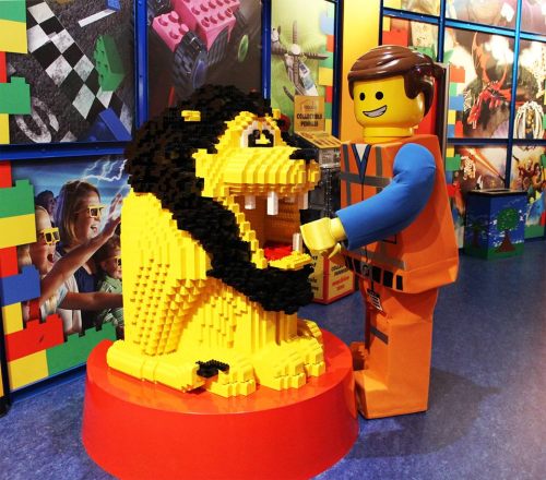 A LEGO figure interacts with a large, yellow LEGO lion sculpture in a colorful play area.