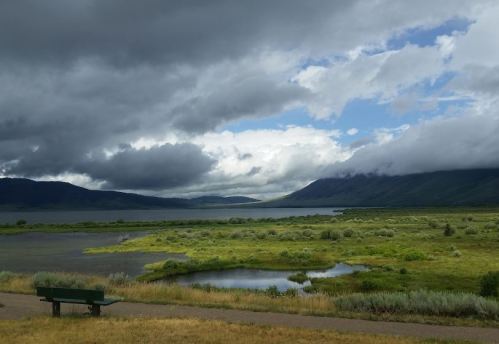 A scenic view of a lake surrounded by green fields and mountains under a cloudy sky, with a bench in the foreground.