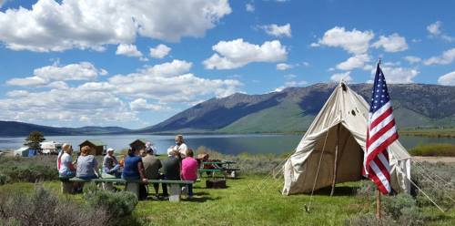 A group of people sits at picnic tables near a tent by a lake, with mountains and an American flag in the background.