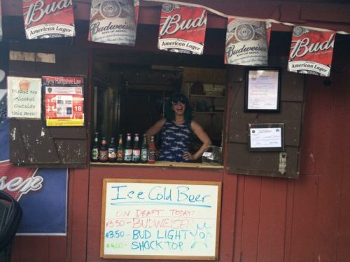 A woman stands behind a bar window with beer options displayed, surrounded by Budweiser banners and a sign for cold beer.