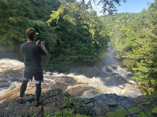 A person stands on a rocky ledge, taking a photo of a rushing river surrounded by lush green trees.