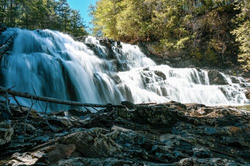 A serene waterfall cascades over rocky terrain, surrounded by lush green trees under a clear blue sky.