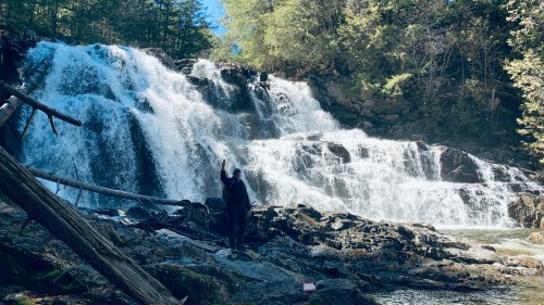 A person stands by a waterfall, raising one hand, surrounded by lush greenery and rocky terrain.