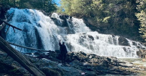 A person stands near a waterfall, raising one hand, surrounded by lush greenery and rocky terrain.