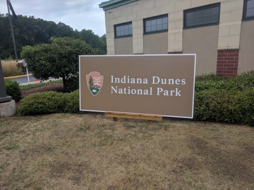 Sign for Indiana Dunes National Park, featuring the park logo and located near a building and landscaped area.
