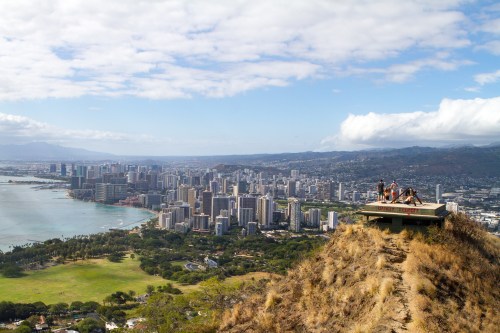 A panoramic view of a city skyline with mountains and ocean, featuring people on a lookout point.