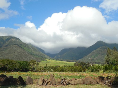 Lush green mountains under a partly cloudy sky, with a field in the foreground and a stone wall.