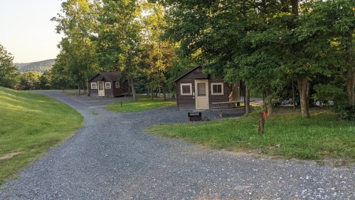 Two rustic cabins along a gravel path, surrounded by trees and greenery, with a scenic hillside in the background.