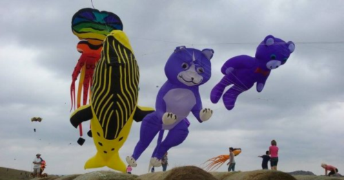Colorful kites in animal shapes, including a bear and a fish, flying against a cloudy sky with people in the background.