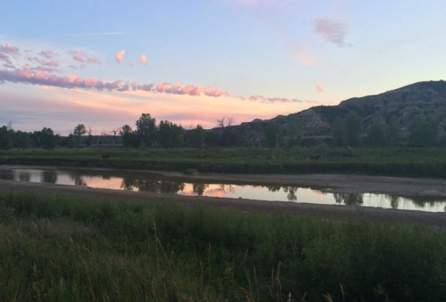 A serene landscape featuring a river reflecting the sky at sunset, surrounded by greenery and distant hills.