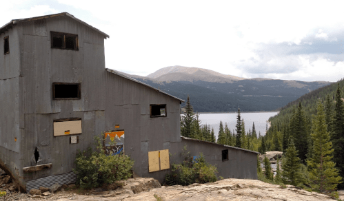 An abandoned building near a lake, surrounded by trees and mountains under a cloudy sky.