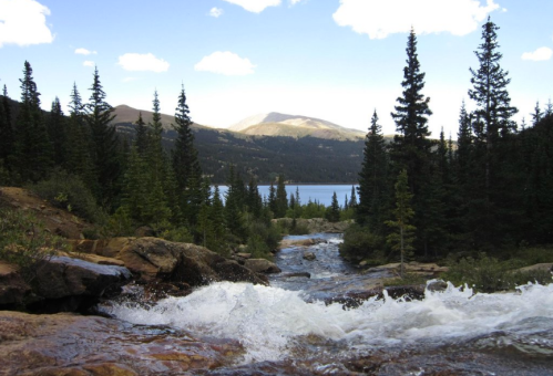 A serene river flows through a forested landscape, with mountains and a lake visible in the background under a blue sky.