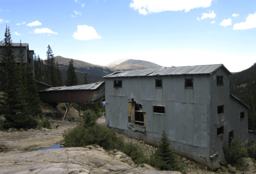 Abandoned gray buildings surrounded by trees and mountains under a clear blue sky.
