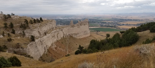 A panoramic view of a rocky cliffside overlooking a valley with fields and distant hills under a cloudy sky.