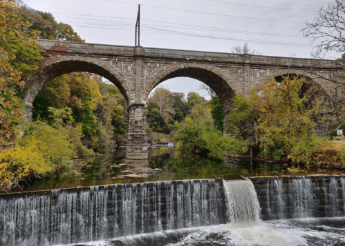 A stone bridge with arches spans over a river, with a waterfall cascading below and autumn foliage surrounding the scene.