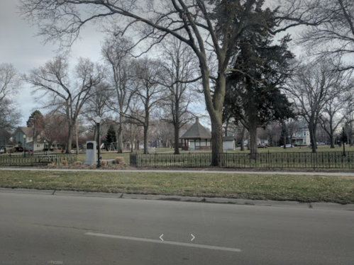 A park scene with bare trees, a gazebo, and a grassy area, viewed from a street.