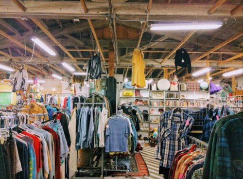 A vintage thrift store interior filled with racks of clothing and various items under a wooden ceiling.