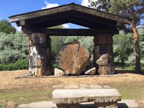 A wooden structure with a large tree stump centerpiece, surrounded by greenery and a stone bench in a park setting.