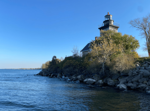 A lighthouse stands on a rocky shore, surrounded by trees, with calm water and a clear blue sky in the background.