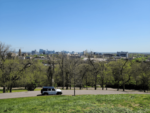 A panoramic view of a city skyline with green trees in the foreground and a clear blue sky. A white vehicle is parked nearby.