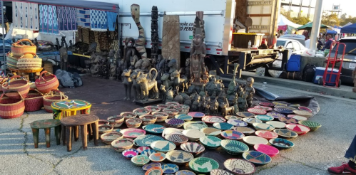 A vibrant market scene featuring colorful woven baskets, carved sculptures, and decorative bowls displayed on the ground.