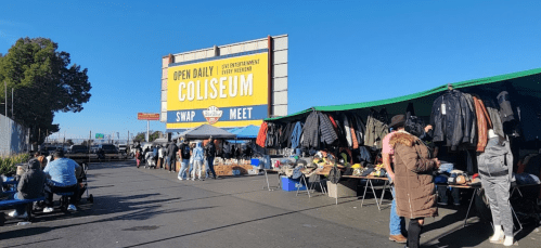 A bustling swap meet outside a coliseum, with vendors selling clothes and various items under colorful tents.