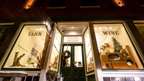 A cozy shop window displaying yarn, wine, and holiday decorations, with warm lighting and a festive wreath.