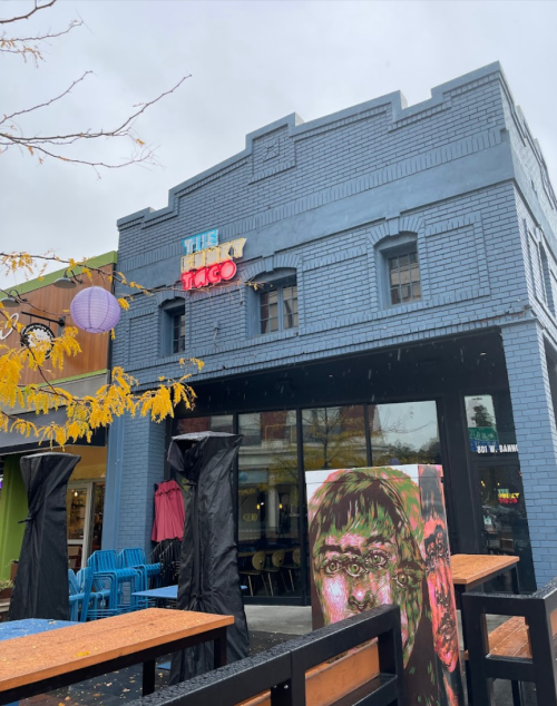 A blue brick building with "The Taco" sign, featuring outdoor seating and a colorful mural of faces.